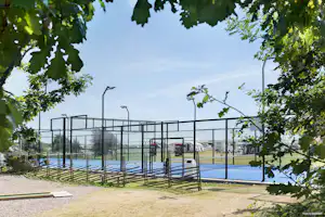 Sportplatz am Campingplatz mit hohem Metallzaun, Sitzbänken und Kunstrasen, im Hintergrund Wiese und Bäume.