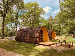 Glamping-Holzhütte mit orangefarbenen Türen und Holzpodest auf einem Campingplatz, umgeben von Bäumen und Rasen.