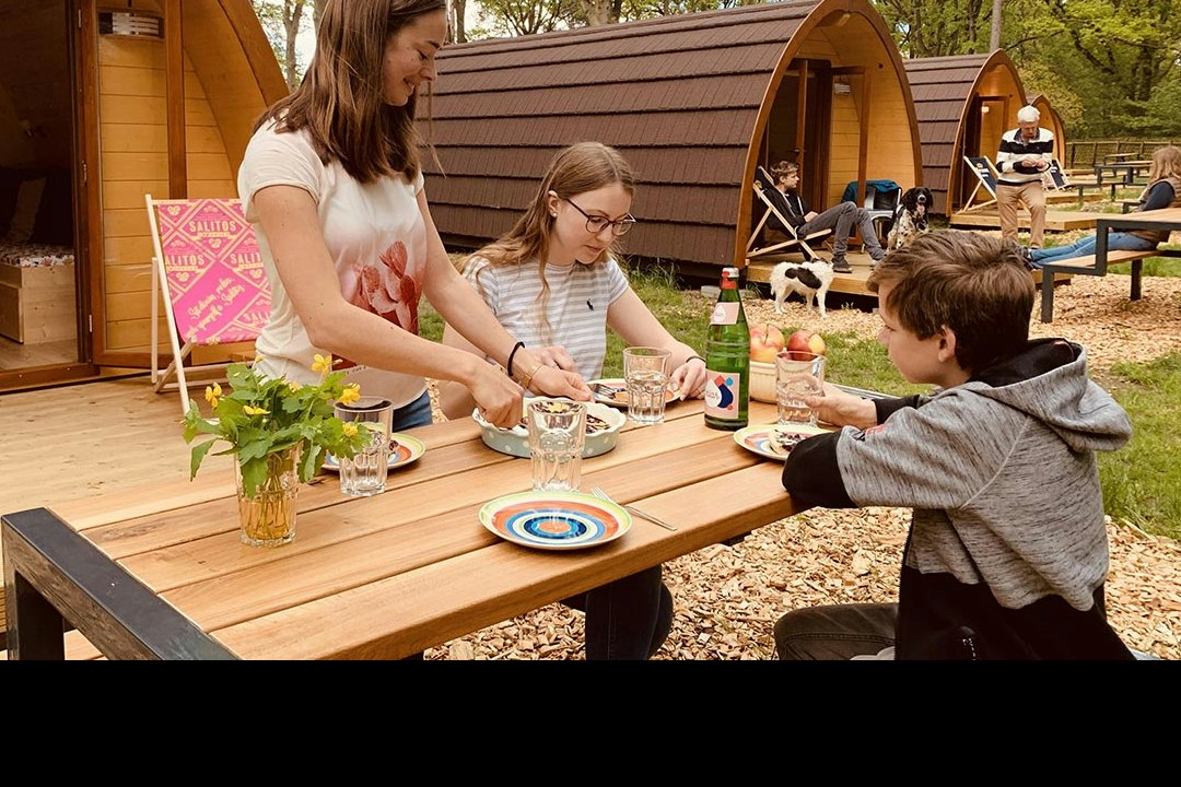 Holzterrasse am Campingplatz mit Tisch, Geschirr und Kindern vor einem Wohnwagen-Zelt, im Hintergrund Bäume und weitere Sitzplätze.