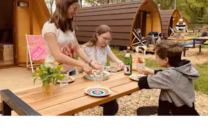 Holzterrasse am Campingplatz mit Tisch, Geschirr und Kindern vor einem Wohnwagen-Zelt, im Hintergrund Bäume und weitere Sitzplätze.