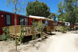Rotes Holzhaus mit Terrasse, Markise und Fenstern, umgeben von Bäumen und Sträuchern auf einem Campingplatz.