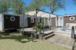 Bungalow mit Holzterrasse, Sonnensegel, Gartenstühlen und Pflanzkübel vor dunkler Holzfassade unter blauem Himmel.
