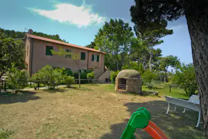 Campinghaus mit Terrasse und Garten, Steinofen im Vordergrund, Rasenfläche und Bäume im Hintergrund.