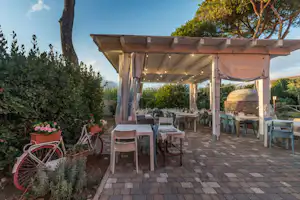 Überdachte Camping-Terrasse mit Tischgarnituren, Stühlen und Lichterkette, gepflasterter Fläche und Pflanzenkübeln im Garten.