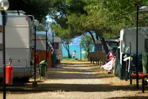 Wohnmobile und Campingfahrzeuge auf einem Kiesweg zwischen Bäumen, mit Mülltonnen und Wäsche am Stellplatz.