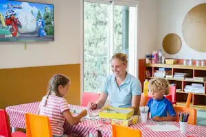 Kinder spielen am Tisch in der Ferienanlage, Betreuerin im blauen Shirt, Fensterblick ins Grüne, Spielzeug und Stifte auf dem Tisch.