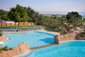 Großes Camping-Schwimmbecken mit mehreren Stufen, Liegen am Rand und Blick auf das Meer hinter Bäumen.