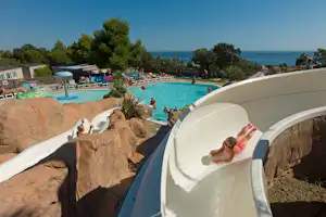 Weiße Wasserrutsche am Pool, daneben Liegen und Bäume, im Hintergrund das Meer und ein helles Campinggelände.