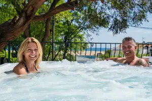 Zwei Personen im Whirlpool eines Campingplatzes, umgeben von Bäumen und Blick auf den Zaun im Hintergrund.