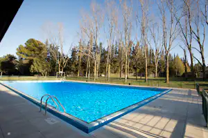 Schwimmbecken auf dem Campingplatz mit gepflasterter Terrasse, Metalltreppe und Bäumen im Hintergrund.