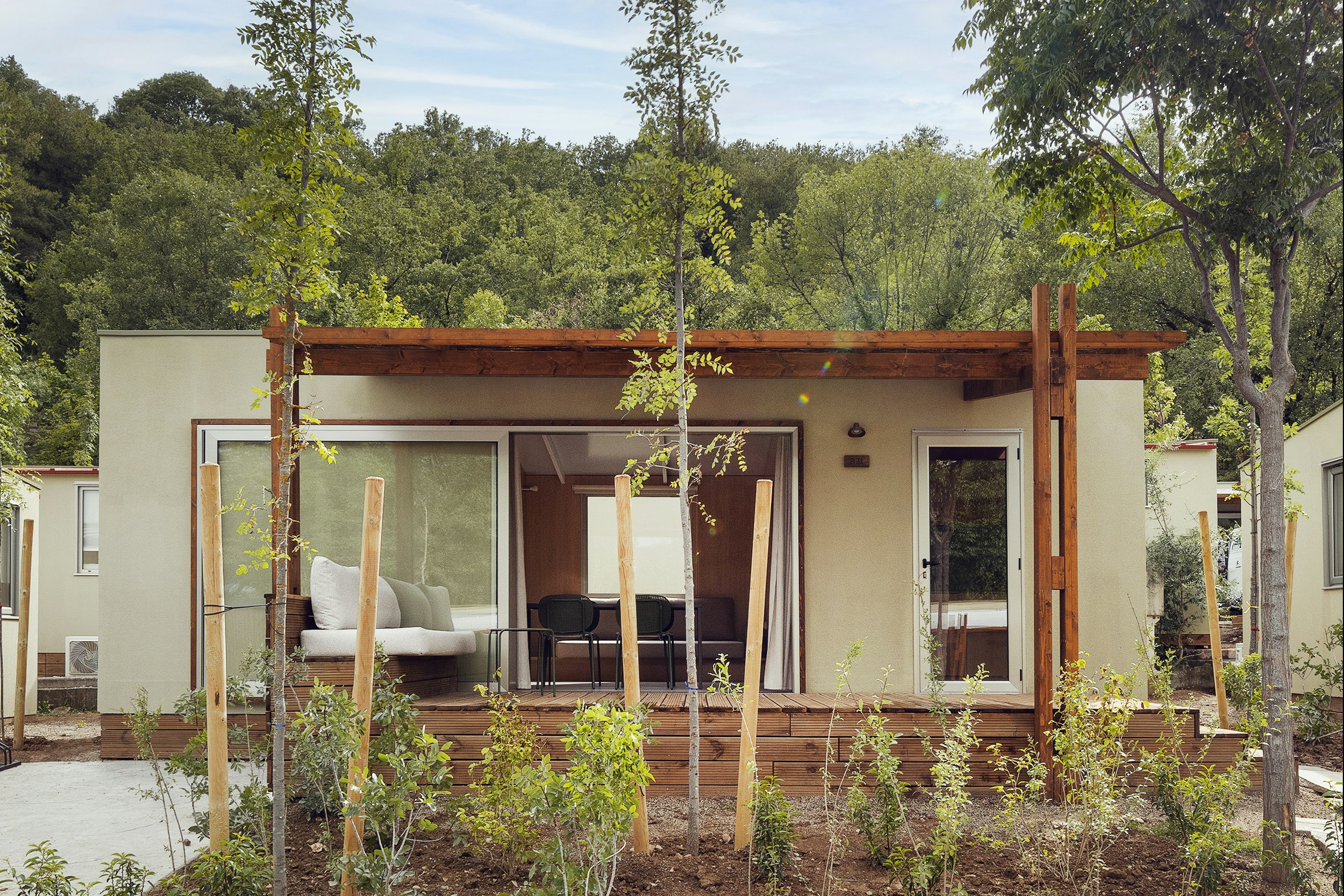 Campinghaus mit Holzpergola und Terrasse, Sitzgruppe vor großen Fenstern, umgeben von jungen Bäumen im Garten.