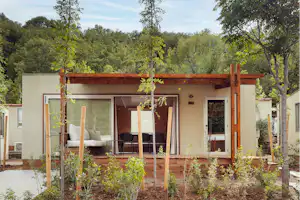 Campinghaus mit Holzpergola und Terrasse, Sitzgruppe vor großen Fenstern, umgeben von jungen Bäumen im Garten.