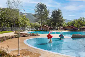Camping-Pool mit Wasserspiel, Sonnendusche und Liegestühlen, dahinter Bungalows und Bäume vor grünen Hügeln.