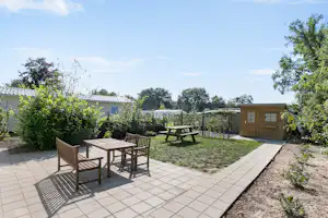 Terrasse mit Tisch und Stühlen, gepflasterter Fläche und Gartenweg, dahinter Gartenhäuser und Zäune, umgeben von Bäumen und Sträuchern.