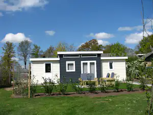 Modernes Mobilheim mit weißen und schwarzen Fassaden, Fenstern und Sitzbereich vor gepflegtem Garten und Zaun.