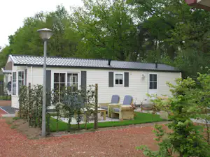 Mobilheim mit Terrasse, Gartenweg und Sitzgruppe vor dem Haus, umgeben von Bäumen und Sträuchern.