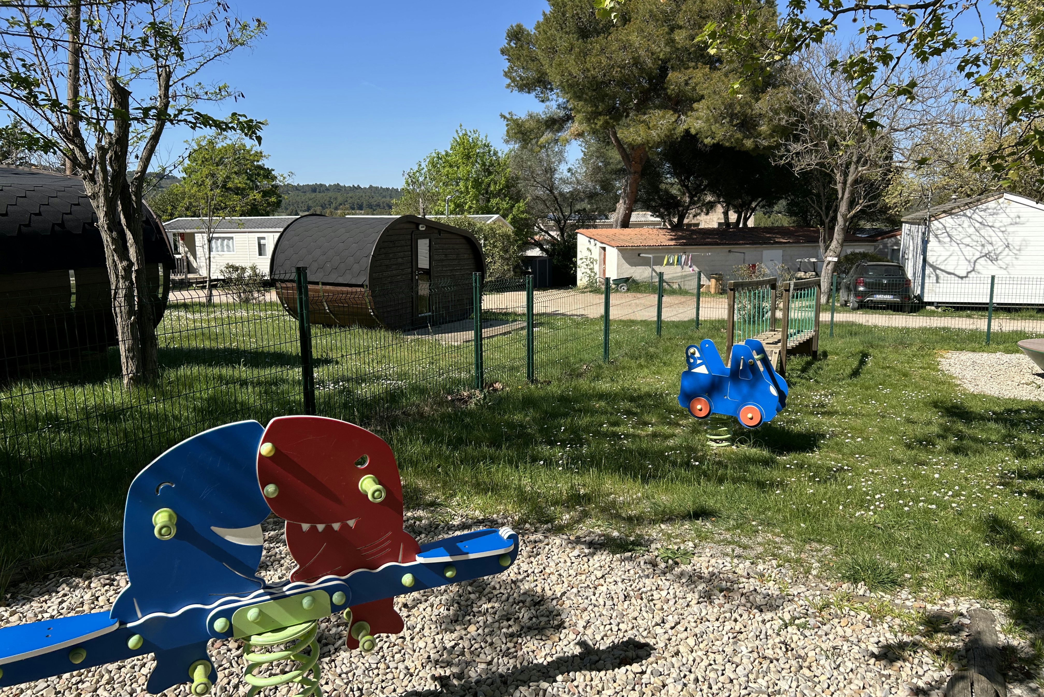 Spielplatz mit blauem Spielgerät und Sandfläche im Campingplatz-Grün, daneben Wohnwagen und Bäume im Hintergrund.