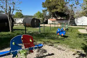 Spielplatz mit blauem Spielgerät und Sandfläche im Campingplatz-Grün, daneben Wohnwagen und Bäume im Hintergrund.