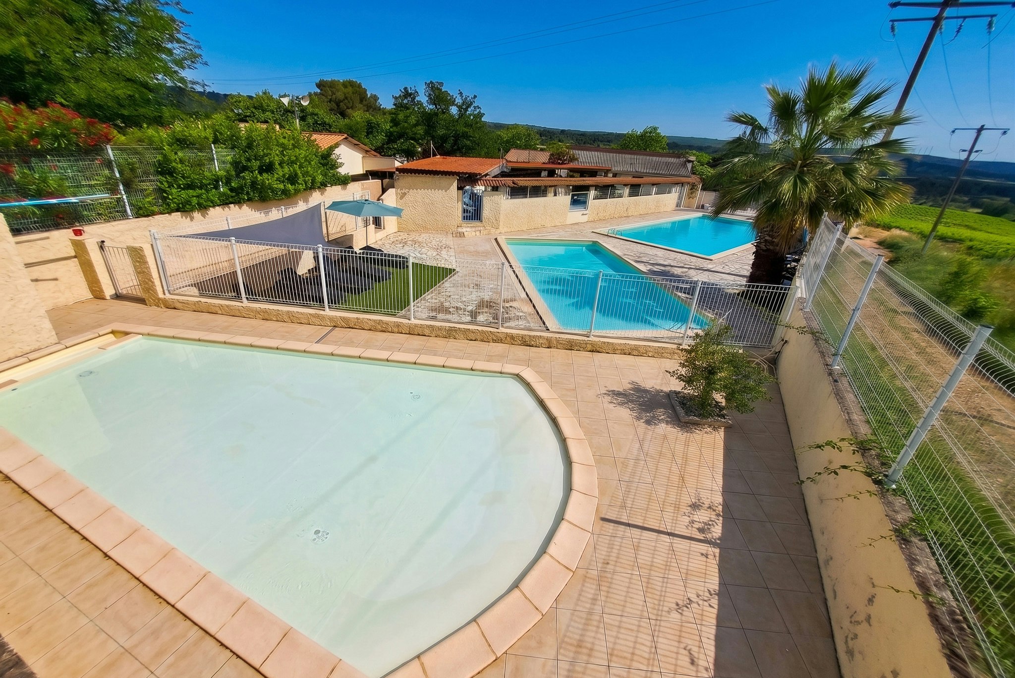 Großer Camping-Pool mit Zaun, daneben ein zweiter Pool, Gebäude und Rasenfläche im Hintergrund unter klarem Himmel.