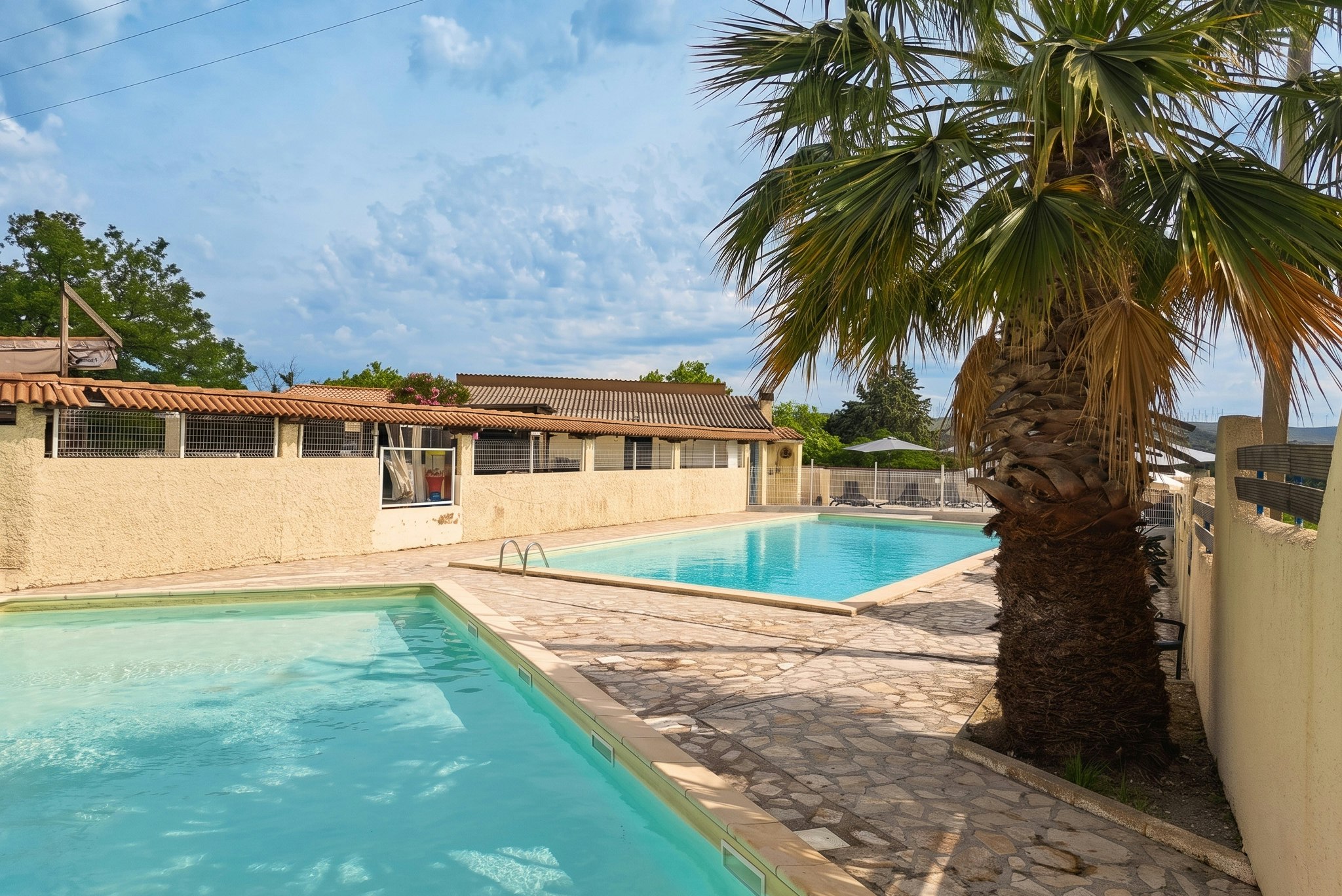 Schwimmbecken und Poolbereich mit Palmen, gepflastertem Weg und Reihen von Bungalows unter blauem Himmel.