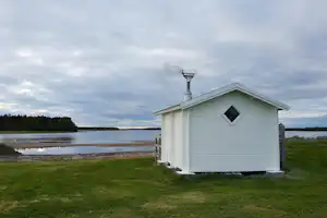 Weißes Saunagebäude am See mit Kamin, Holzsteg und Uferlinie im Hintergrund, umgeben von Bäumen.