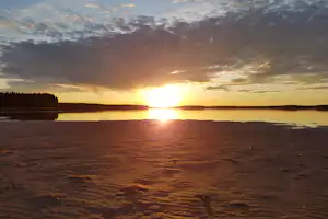 Sonnenuntergang am See mit spiegelndem Licht auf dem Wasser, sandigem Ufer und Waldlinie am Horizont.