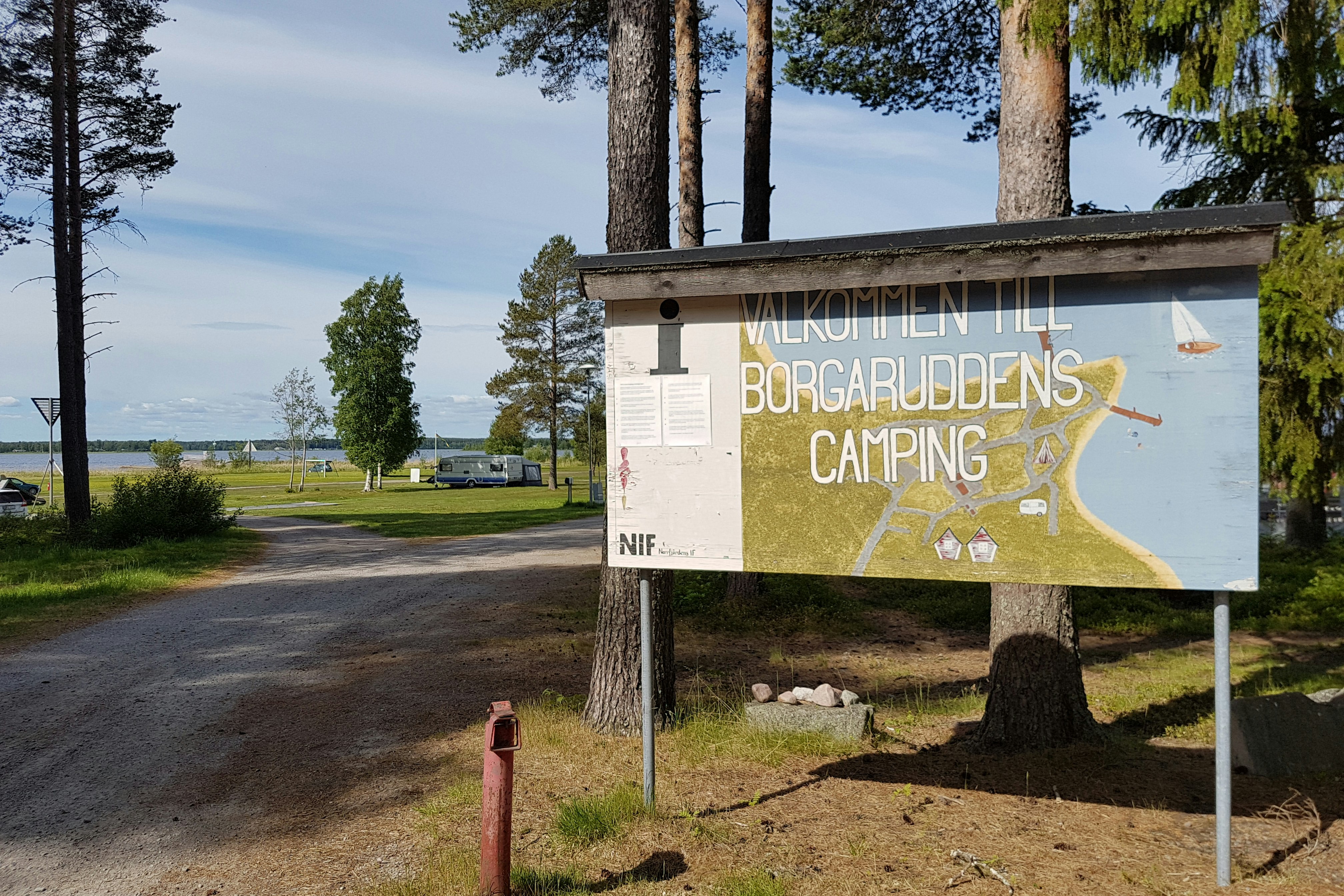 Camping Borgaruddens Havsbad - Willkommensschild und Einfahrt zum Campingplatz im Vordergrund, Wohnwagen- und Zeltstellplatz mit Blick aufs Meer im Hintergrund