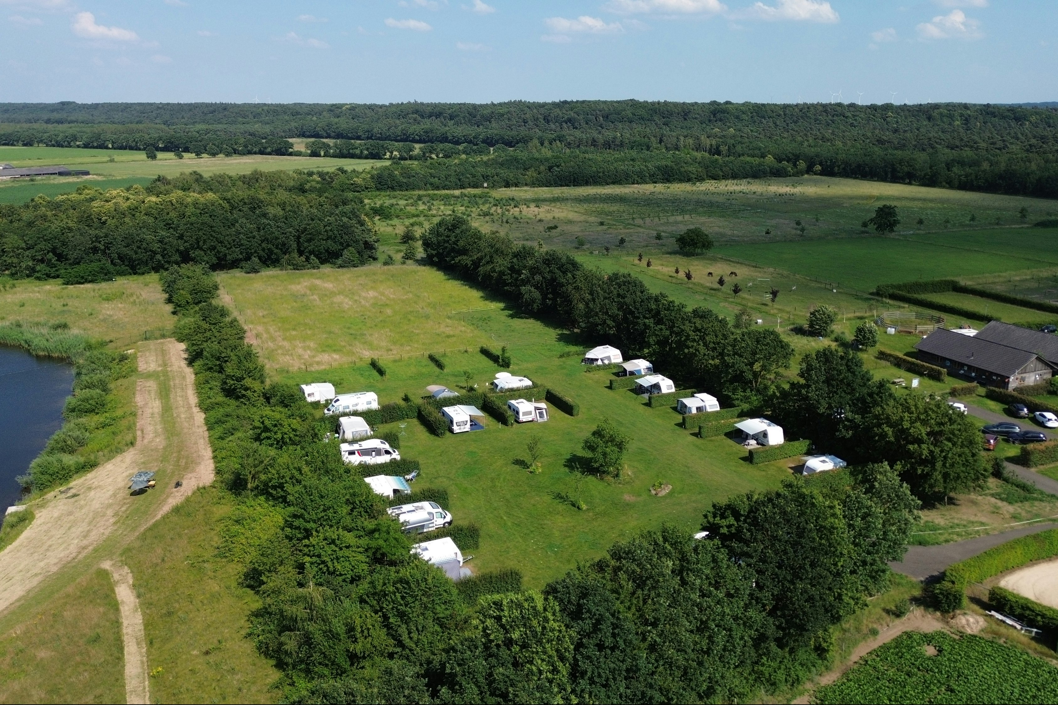 Luftaufnahme eines Campingplatzes mit mehreren Wohnwagen und Zelten auf einer Wiese, am Rand ein See und Wege.