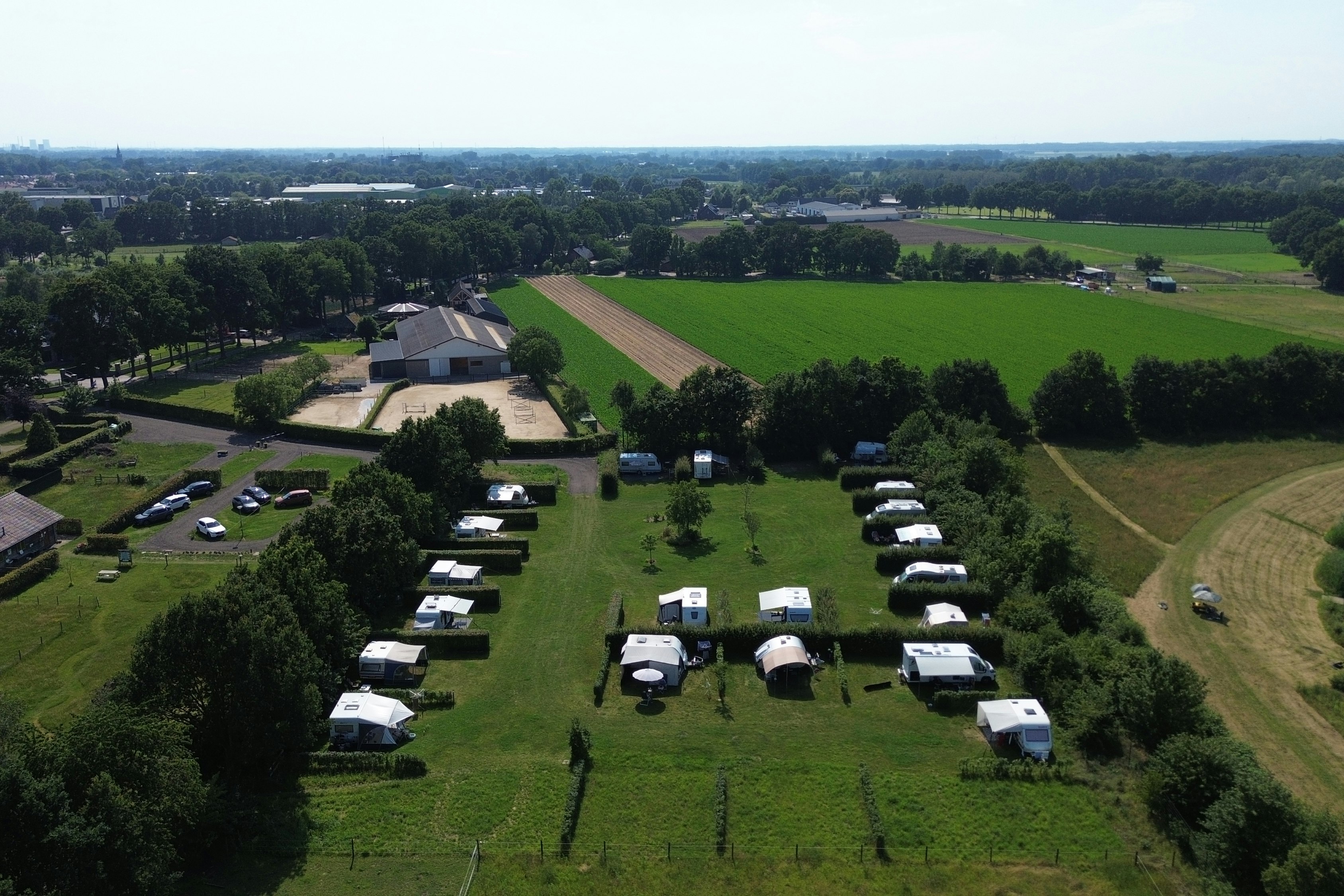 Luftaufnahme eines Campingplatzes mit mehreren Stellplätzen, Wohnwagen und Zelten, umgeben von Feldern, Wegen und Bäumen.