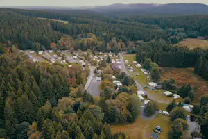 Luftaufnahme eines Campingplatzes im Wald mit Stellplätzen, kleinen Gebäuden und Zufahrtsstraße zwischen Bäumen.