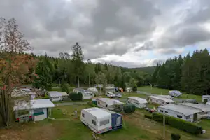 Campingplatz mit Wohnwagen und kleinen Gebäuden, umgeben von Bäumen und Hecken, im Hintergrund Hügel und bewölkter Himmel.