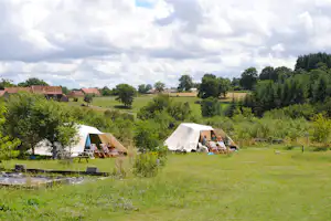 Zelte auf einem Campingplatz auf einer grünen Wiese, mit Sitzgelegenheiten und Bäumen im Hintergrund, Blick über die Landschaft.