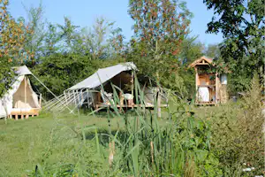 Campingzelte im Grünen, mit Rasenfläche und Bäumen im Hintergrund, daneben ein Holz-Spielturm und ein kleiner Holzkasten.