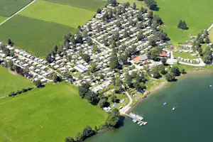 Luftaufnahme eines Campingplatzes am See mit vielen Stellplätzen, Bäumen, Wegen und Bootssteg am Ufer.