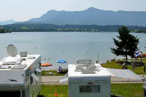 Wohnwagen am Campingplatz am Seeufer, mit Sonnenschirmen, Rasenfläche und Blick auf Berge und Bäume.