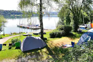 Zelt auf einem Campingplatz am See, mit Picknicktisch, Bäumen und Blick auf Segelboote im Wasser.