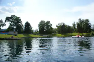 Campingsee mit Schwimmsteg, Ruderbooten und aufblasbarem Spielzeug am Ufer, dahinter Rasen und Bäume.