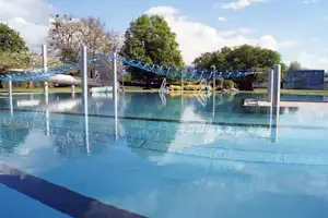 Schwimmbecken mit Sicherheitsnetz, Sprungstangen und Wasserfontänen, im Hintergrund Bäume und ein Gebäude am Campingplatz.