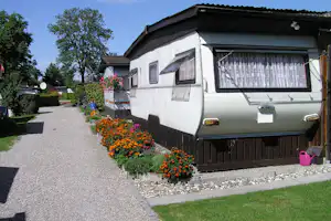 Wohnwagen mit Markise und Vorhangfenstern, Blumenbeet am Stellplatz und Kiesweg zwischen Bäumen.