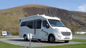 Weißes Wohnmobil auf einem Stellplatz mit Stromanschluss, im Hintergrund Berge und ein bewachsener Hügel.