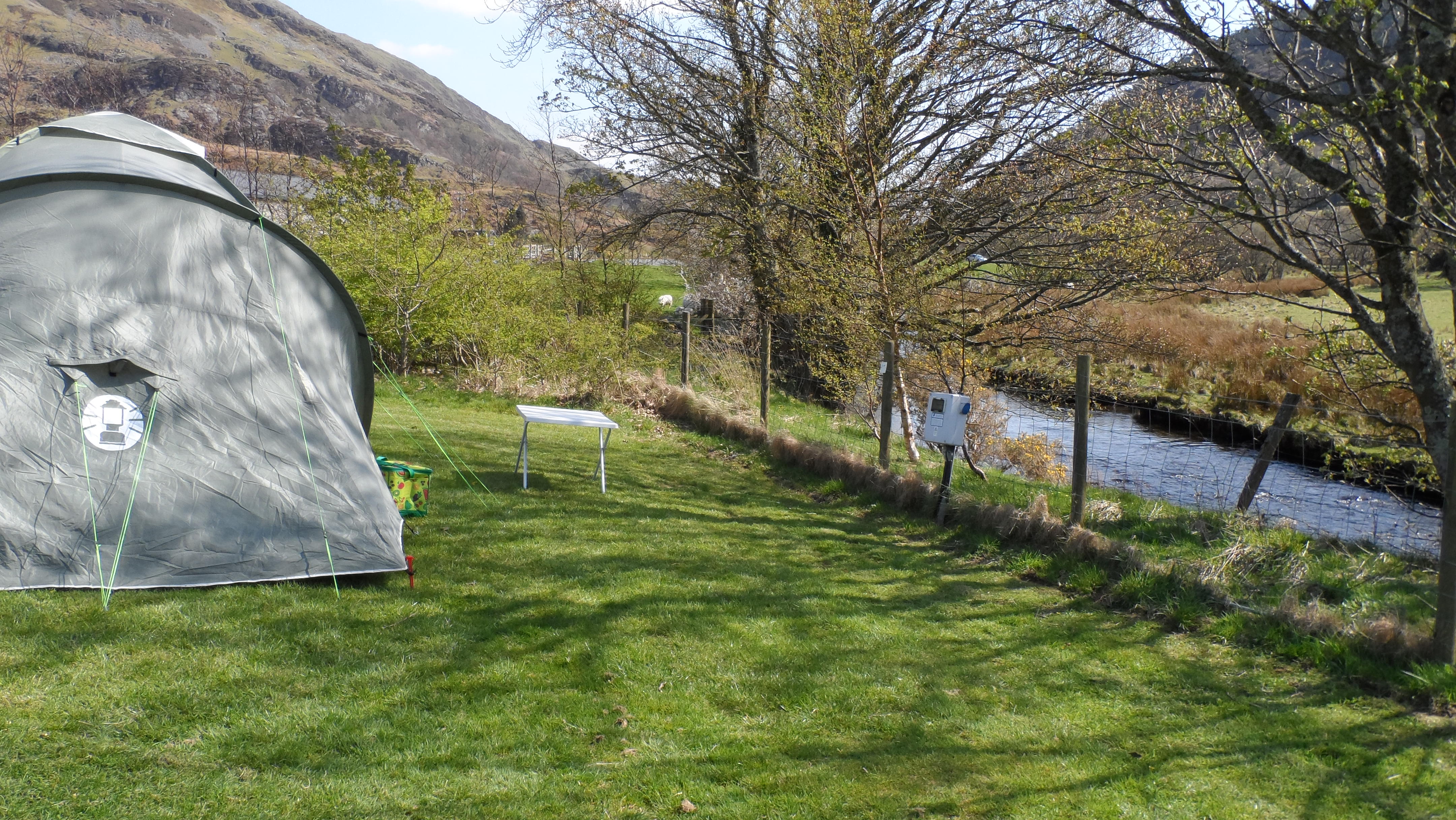 Campingzelt auf einer grünen Wiese am Fluss, im Hintergrund Berge und Bäume, daneben ein weiterer Zeltplatz.