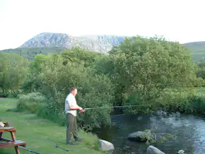 Angler am Flussufer mit Angelrute, daneben Picknicktisch, grünes Ufer und Bäume vor bewaldeten Bergen.