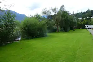 Grünes Campinggrundstück mit Rasenfläche, Bäumen und Hecke, im Hintergrund ein Wohnwagen und Berge.
