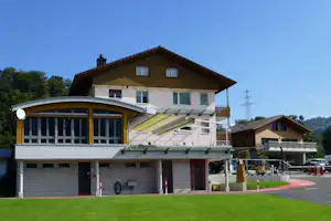 Campinggebäude mit Holzfassade, großer Markise und Terrasse, daneben Rasenfläche und Parkplatz mit Autos.
