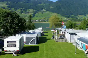 Wohnwagen und Zelte auf einer grünen Stellfläche am See, mit Campingfahrzeugen, Flagge und Blick auf die Berge.