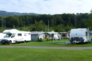Zwei Wohnmobile auf einem Campingplatz mit grünem Gras, Vorzelten, Stühlen und Tischen vor Waldhügeln.