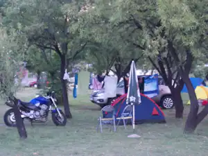 Campingzelt auf einem Grasplatz zwischen Bäumen, daneben Stühle und ein Motorrad, im Hintergrund weitere Zelte und Fahrzeuge.