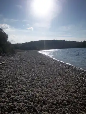 Kiesstrand am Fluss mit ruhigem Wasser, Uferlinie und bewaldeten Hügeln in der Ferne.