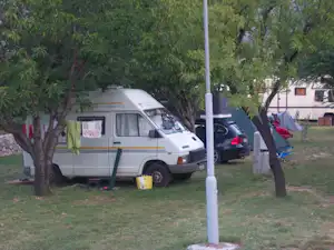 Wohnmobil auf einem Stellplatz im Grünen, mit Grasfläche, Bäumen und einem geparkten Auto im Hintergrund.
