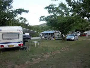 Wohnwagen auf einem Campingplatz mit Vorzelt, daneben Tisch und Stühle, Auto und weitere Wohnwagen im Gras zwischen Bäumen.